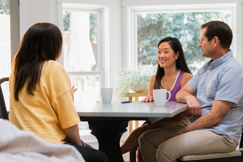 Three people are sitting at a table with coffee mugs, engaged in a lively discussion about childcare tips in a bright room. They seem to be focusing on 5 things to know before hiring a nanny, sharing insights and experiences over their warm drinks.