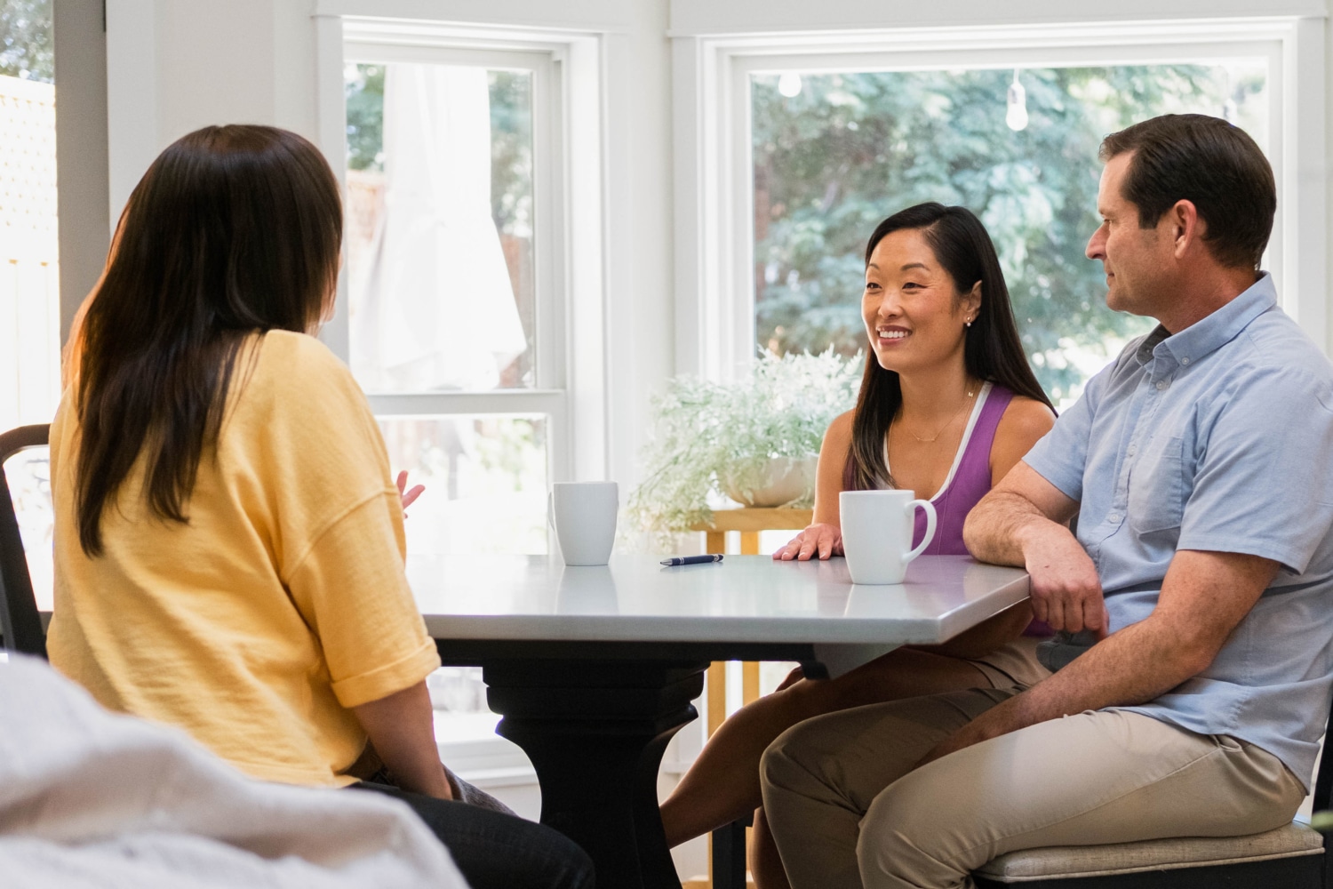 Three people are sitting at a table with coffee mugs, engaged in a lively discussion about childcare tips in a bright room. They seem to be focusing on 5 things to know before hiring a nanny, sharing insights and experiences over their warm drinks.