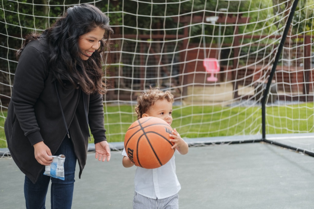A woman smiles at a child holding a basketball in front of a soccer net outdoors, sparking creative learning activity ideas for nannies to engage kids in multi-sport fun.