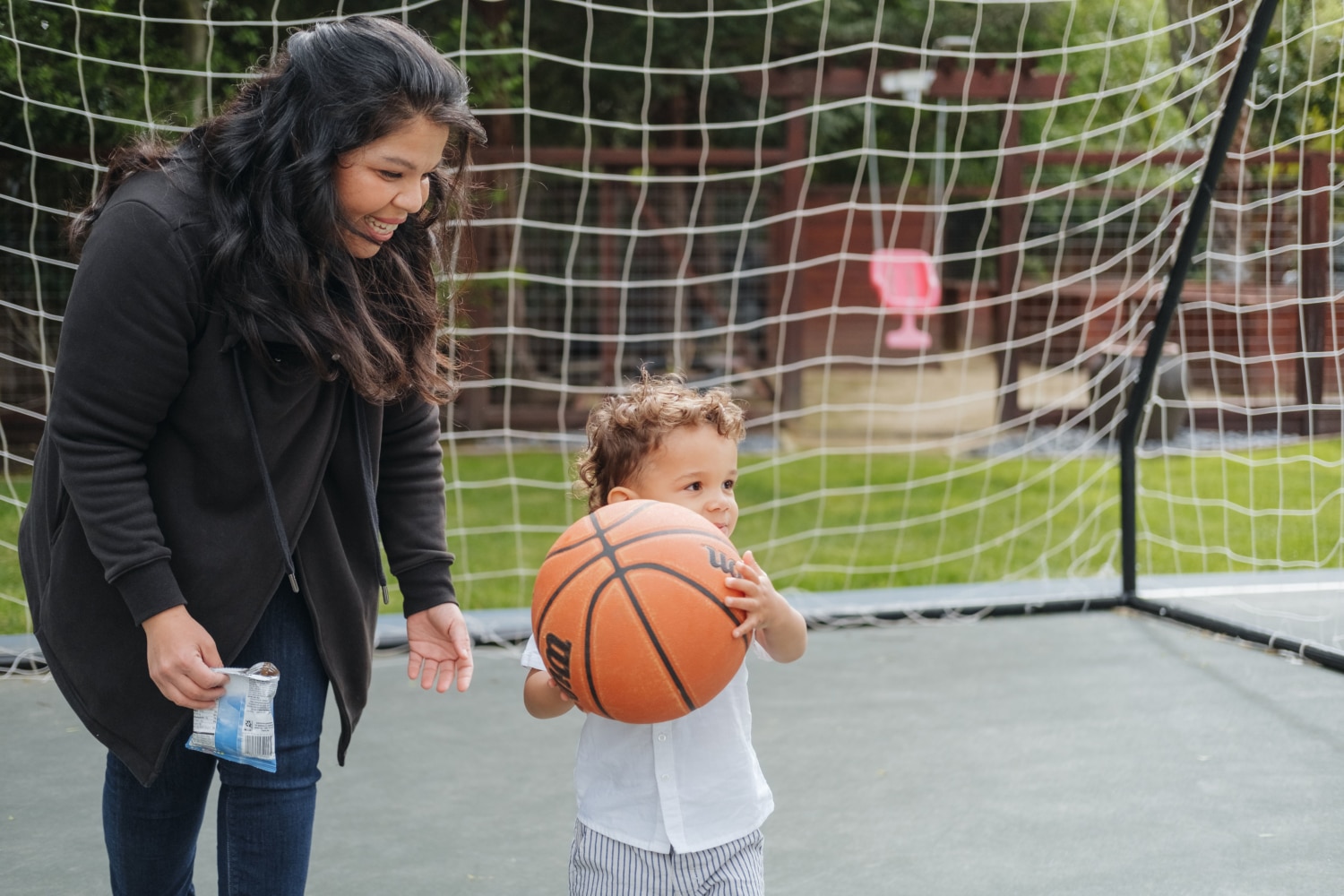 A woman smiles at a child holding a basketball in front of a soccer net outdoors, sparking creative learning activity ideas for nannies to engage kids in multi-sport fun.