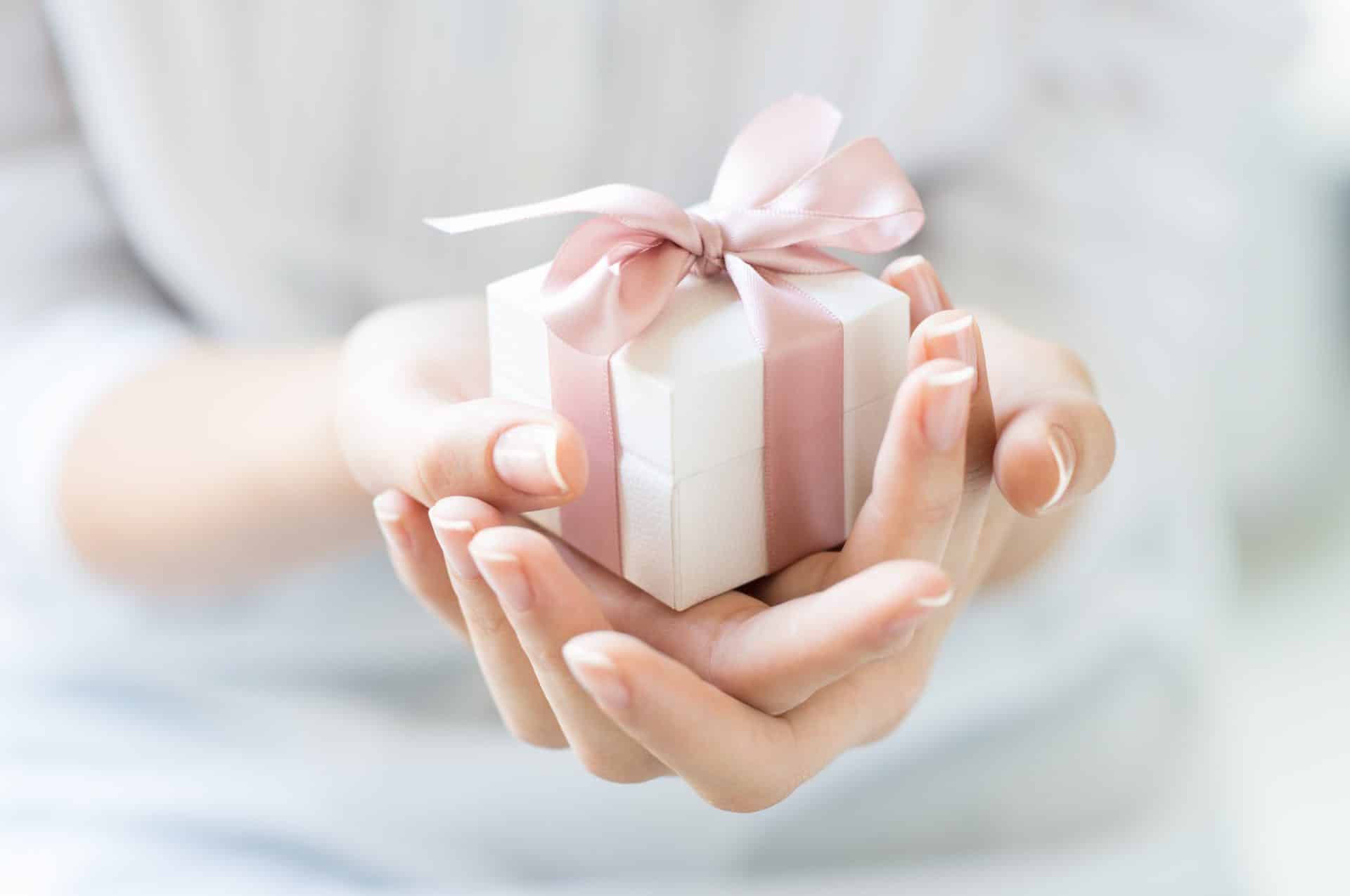 Hands gently cradle a small white gift box adorned with a pink ribbon, perfect as a thoughtful Christmas gift for your nanny.