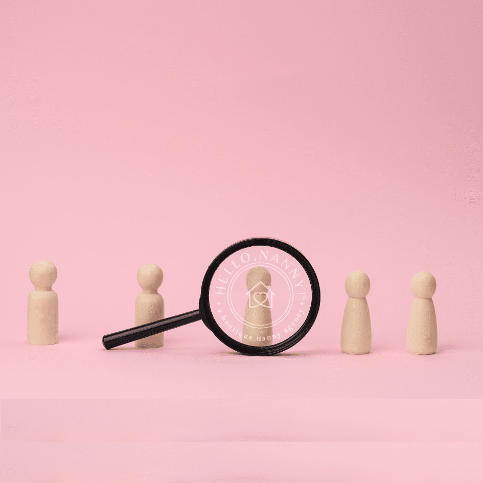 Wooden figures and a magnifying glass with a logo on a pink background illustrate the meticulous process of learning how to screen a nanny effectively.