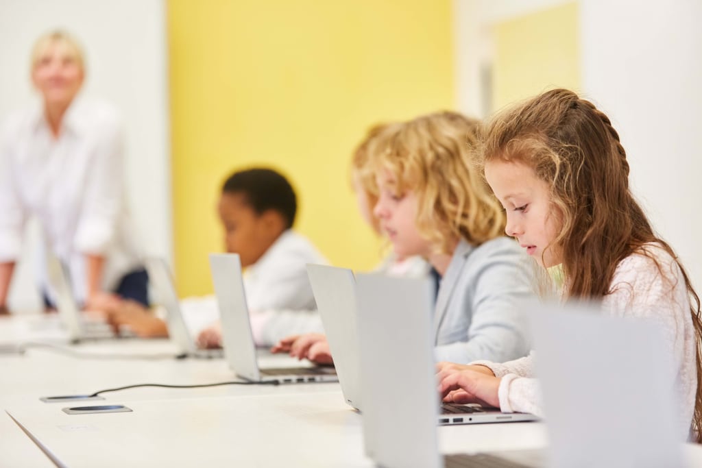 Children focused on laptops in a classroom, with a teacher in the background near a yellow wall.