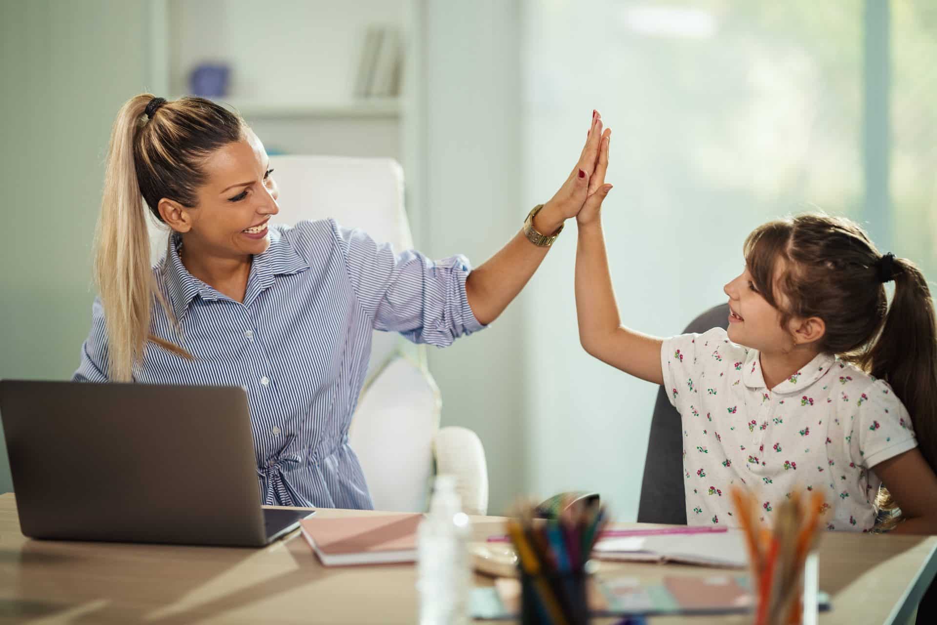 Woman and girl smiling and sharing a high-five at a desk with a laptop and colorful stationery, guided by an experienced nanny.