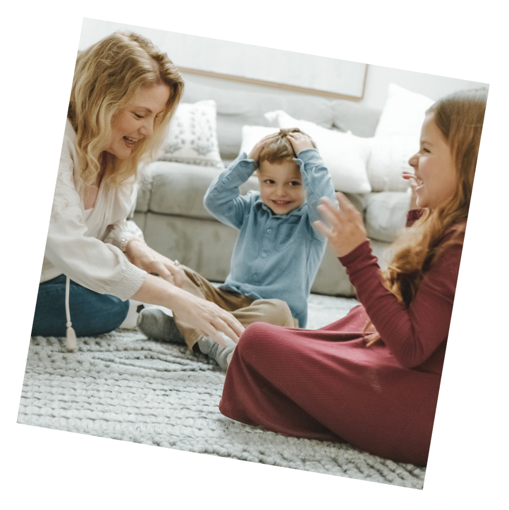 A live-out nanny plays on the floor with a young boy and girl, all smiling and sitting on a patterned rug in a cozy room.