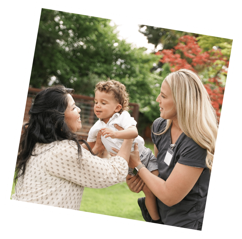 Two women smiling and holding a toddler outdoors, surrounded by green trees and a sunny sky, enjoying a day out with their live-out nanny.