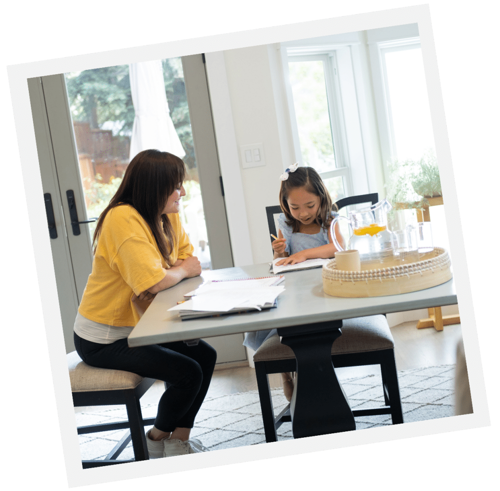 A woman and child sit at a table, reading and writing together in a bright room, creating an atmosphere reminiscent of time spent with a dedicated live-out nanny.