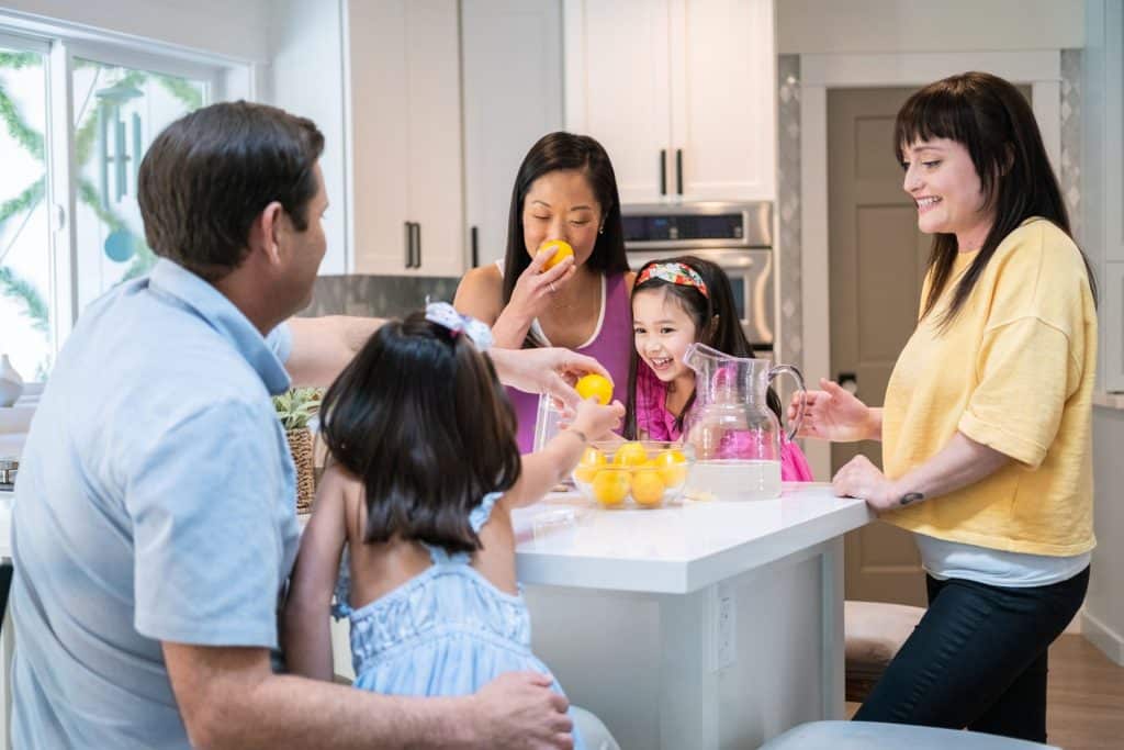 A family of five, along with their live-out nanny, enjoys oranges around a kitchen island, with a pitcher of refreshing lemonade at the center.