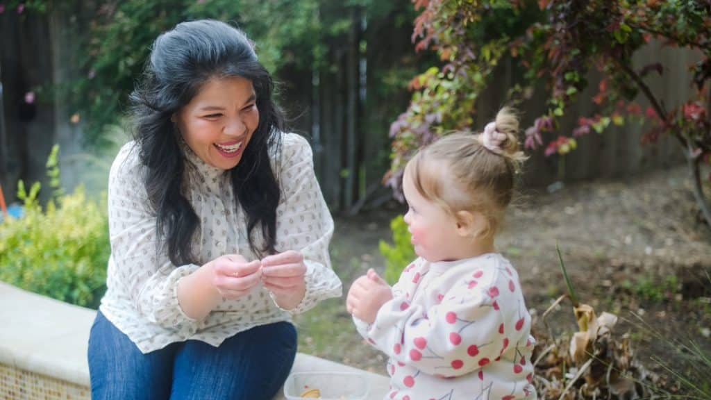 A woman and a child sit outdoors, smiling and interacting near some plants.