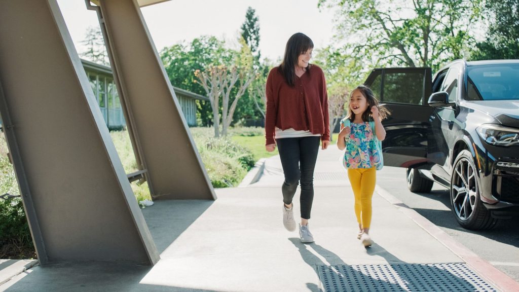 A woman and a young girl joyfully walk hand in hand on a sunny day, with their live-out nanny keeping a watchful eye near the parked car.