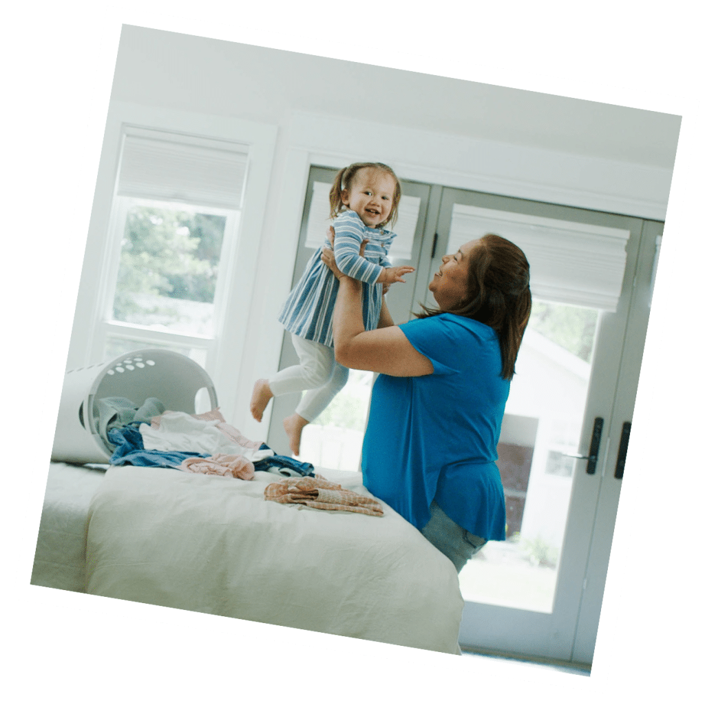 A live-in nanny lifts a smiling toddler near a bed with laundry in a bright room with large windows.