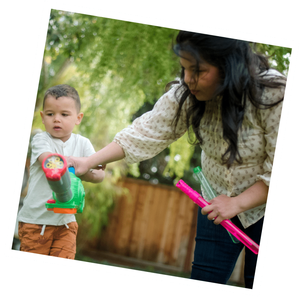 In the garden, a woman and child joyfully play with water guns amidst lush greenery, their laughter echoing through the trees. Watching over them, their live-in nanny ensures everything is safe and fun.