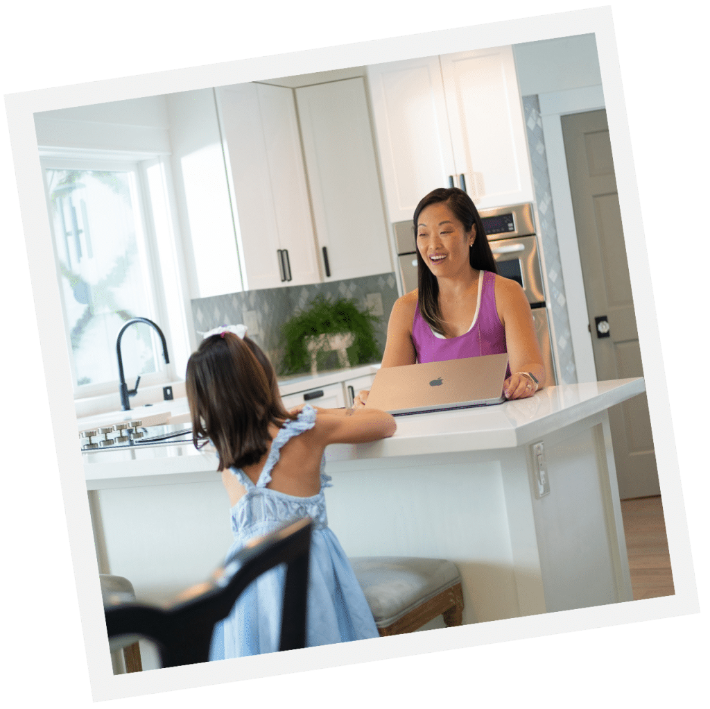 A woman and a child in a blue dress smile and talk at the kitchen counter with their live-in nanny nearby, while a laptop sits open, joining in their lively conversation.