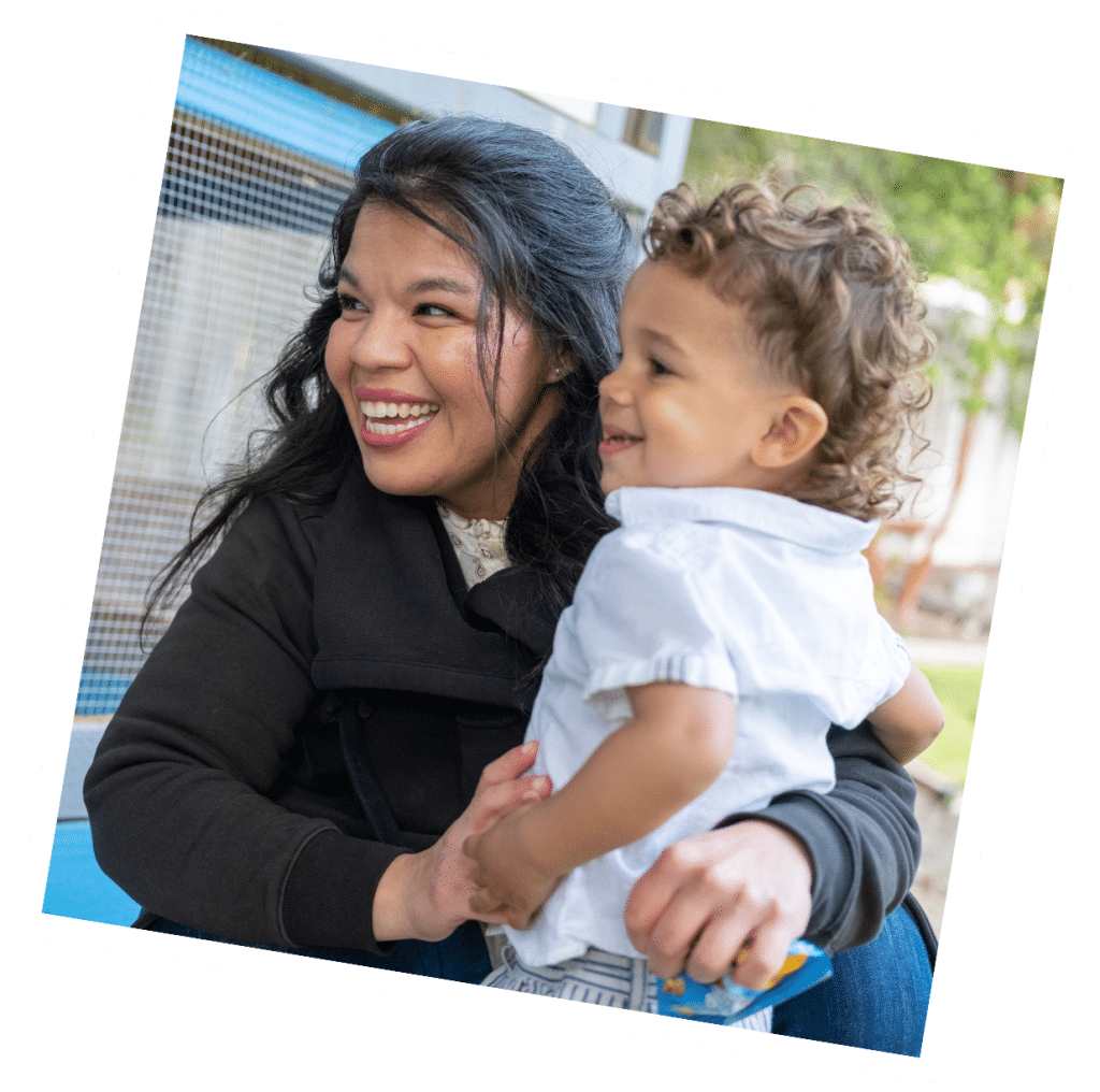 A smiling woman, enjoying her role as a part-time nanny, cradles a happy toddler while sitting outdoors near a fence.