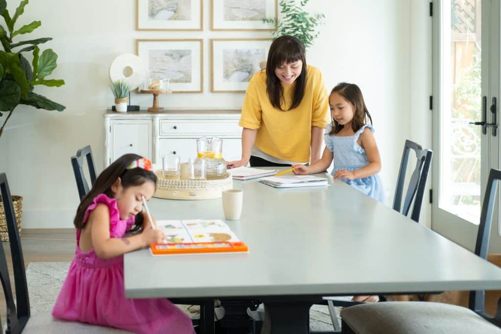 A live-in nanny assists two young girls with their homework at the dining table in a bright, modern room.