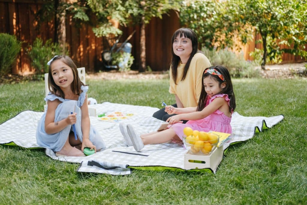 Three people enjoy a picnic on a blanket in a backyard with lush trees and grass. As they relax, their part-time nanny keeps an eye on the playful kids nearby. A basket of lemons sits invitingly within reach, adding a zestful touch to the serene scene.