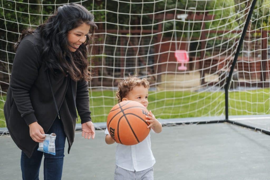 A live-in nanny smiles warmly as the toddler she cares for holds a basketball in front of an outdoor net, capturing a joyful moment in their daily adventures.