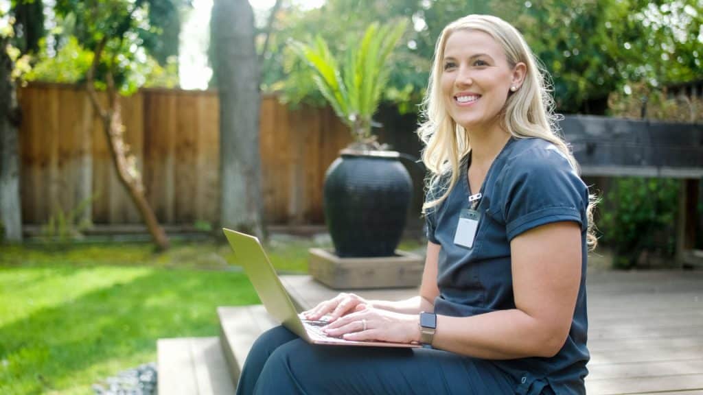 A woman in scrubs, perhaps a dedicated homeschool nanny, types diligently on her laptop outdoors, sitting comfortably on a wooden step with lush greenery providing a serene backdrop.