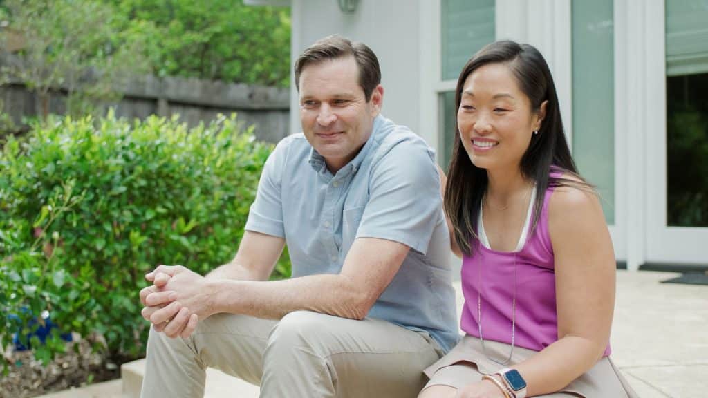A smiling couple sits outdoors on steps, surrounded by lush greenery, with a building in the background. They enjoy a peaceful moment, their live-in nanny taking care of chores inside.