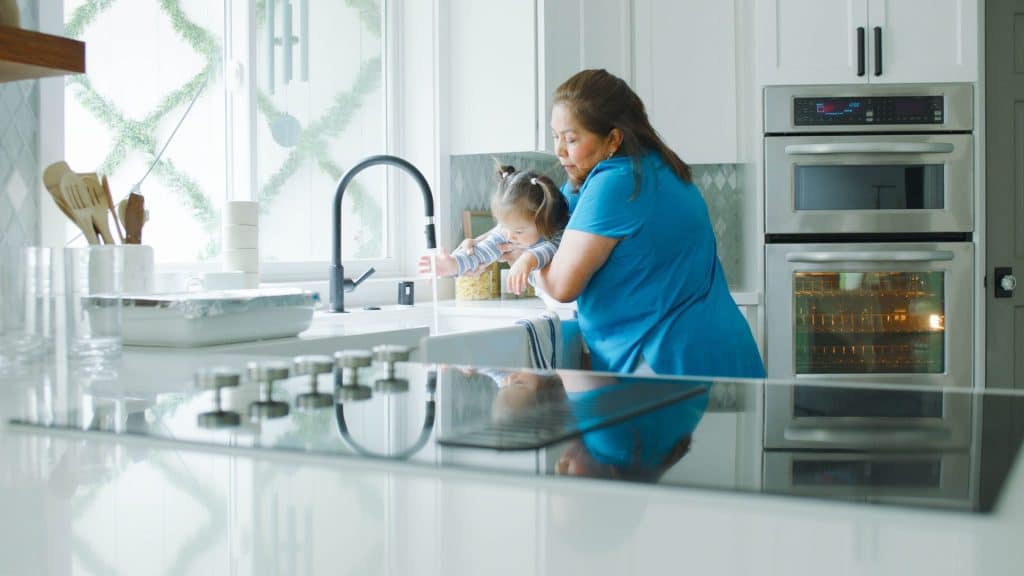 A part-time nanny in a blue shirt helps a child wash hands in a bright kitchen with modern oven and white cabinets.
