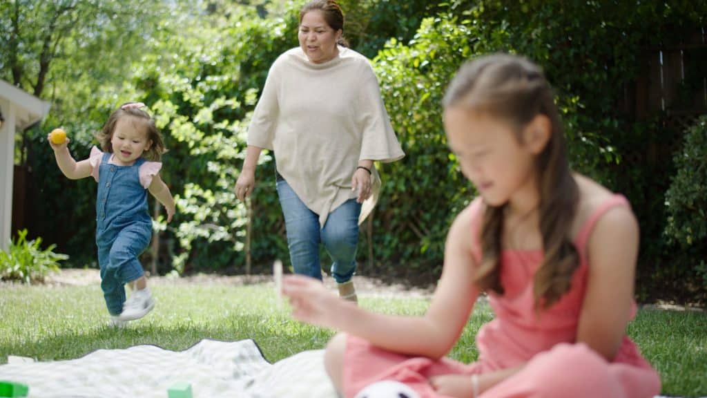 A young girl runs joyfully with a ball in a garden while her live-in nanny watches. Another child plays on the grass nearby.