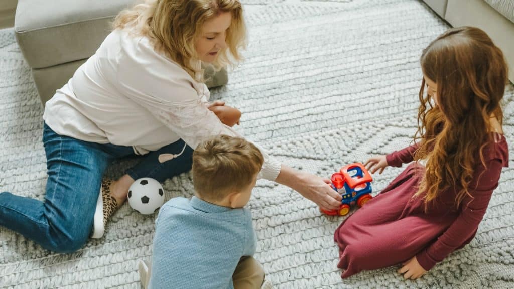An adult and two children, watched over by their attentive part-time nanny, sit on a carpet playing with a toy train while a soccer ball rests nearby.
