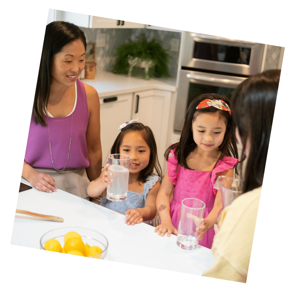 In a cheerful kitchen scene, two children sip water under the watchful eye of a caring adult. A vibrant bowl of lemons sits invitingly on the counter, highlighting a nurturing environment that's perfect for those considering Nanny Consulting Services to elevate their family dynamic.