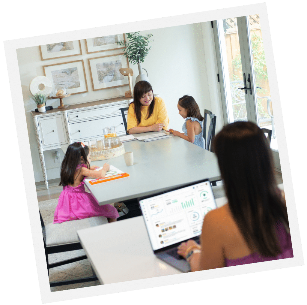 A woman is busy working on a laptop at a table, while another woman, perhaps from a nanny consulting service, engages in lively conversation with two children at a nearby dining table.