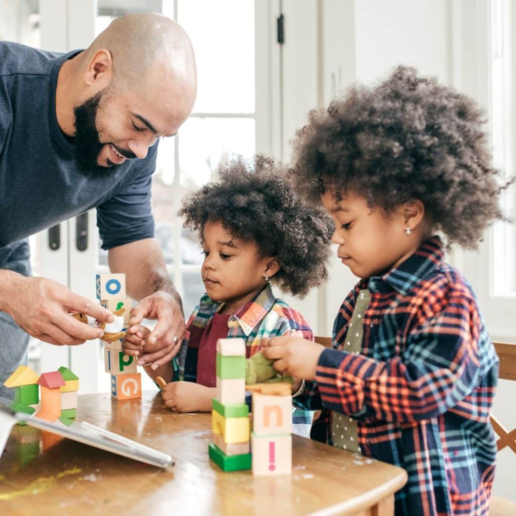 A father and two young children engage in early childhood development as they build with colorful blocks at a table in a bright room.