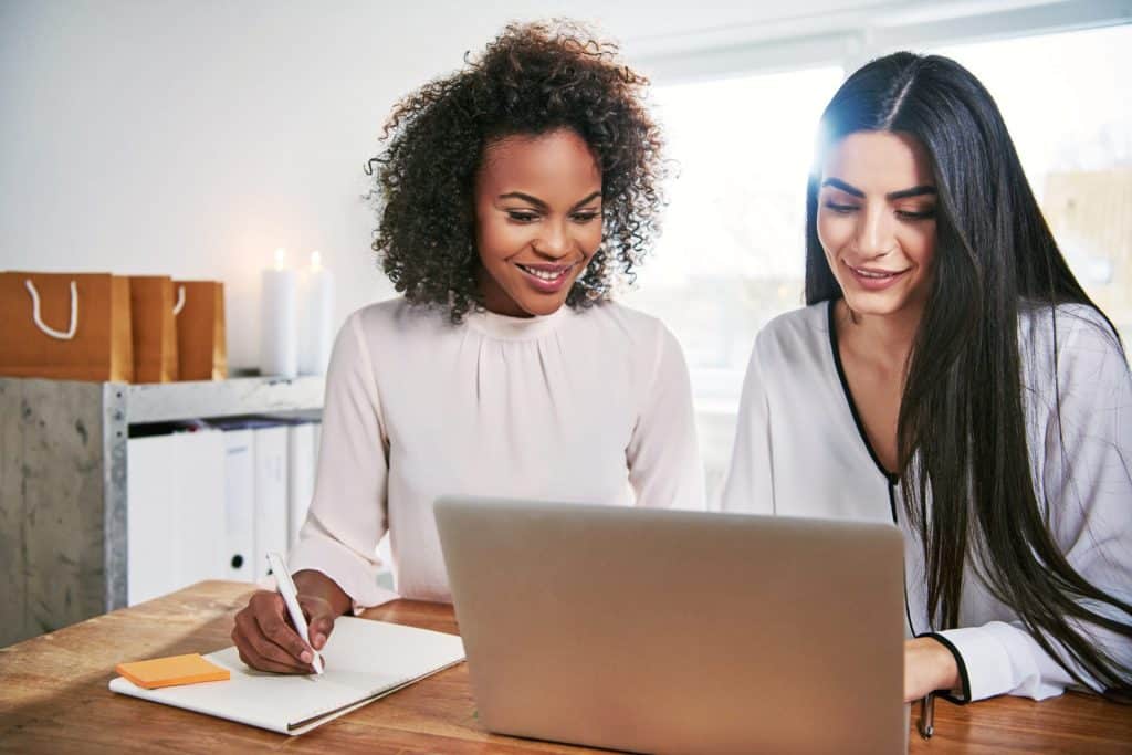Two women smiling and collaborating at a desk, with one taking notes while both focus on a laptop screen, reviewing Nanny Consulting Services.