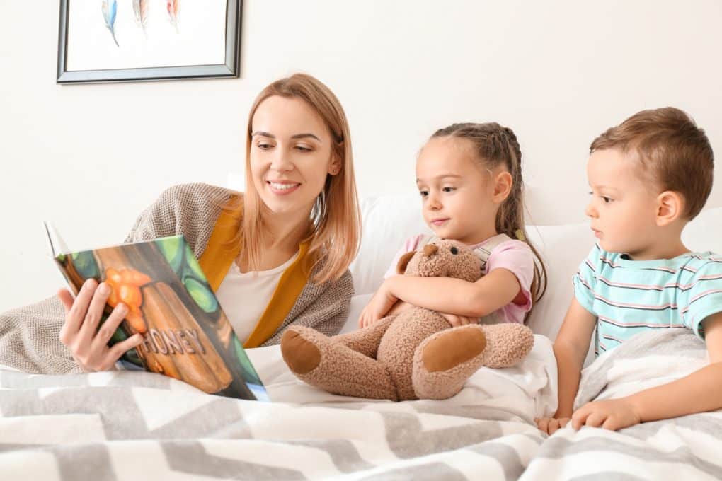 A woman reads a bedtime story to two children nestled in bed; one child clutches a teddy bear, offering comfort much like learning how to have a tough conversation with a nanny can ease parental concerns.