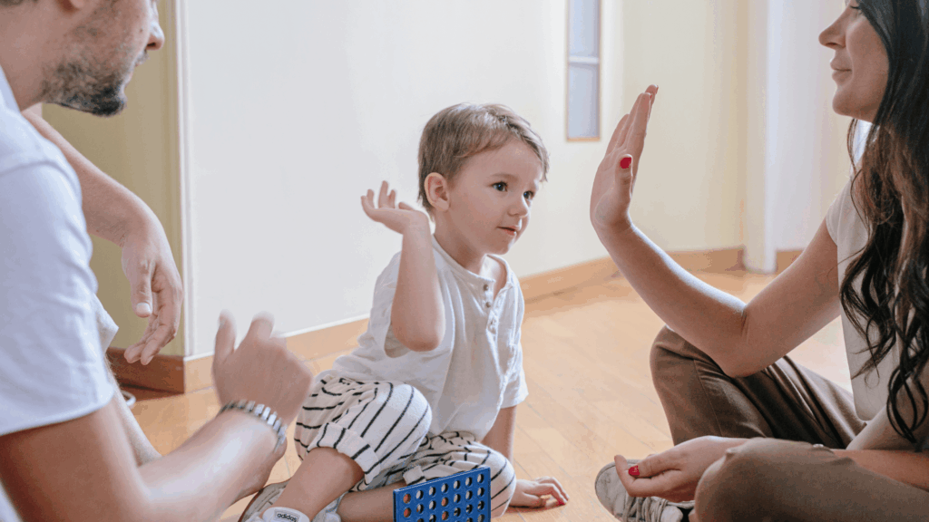 Default parent interacting with Small child sitting on the floor, giving a high-five, with another adult nearby.