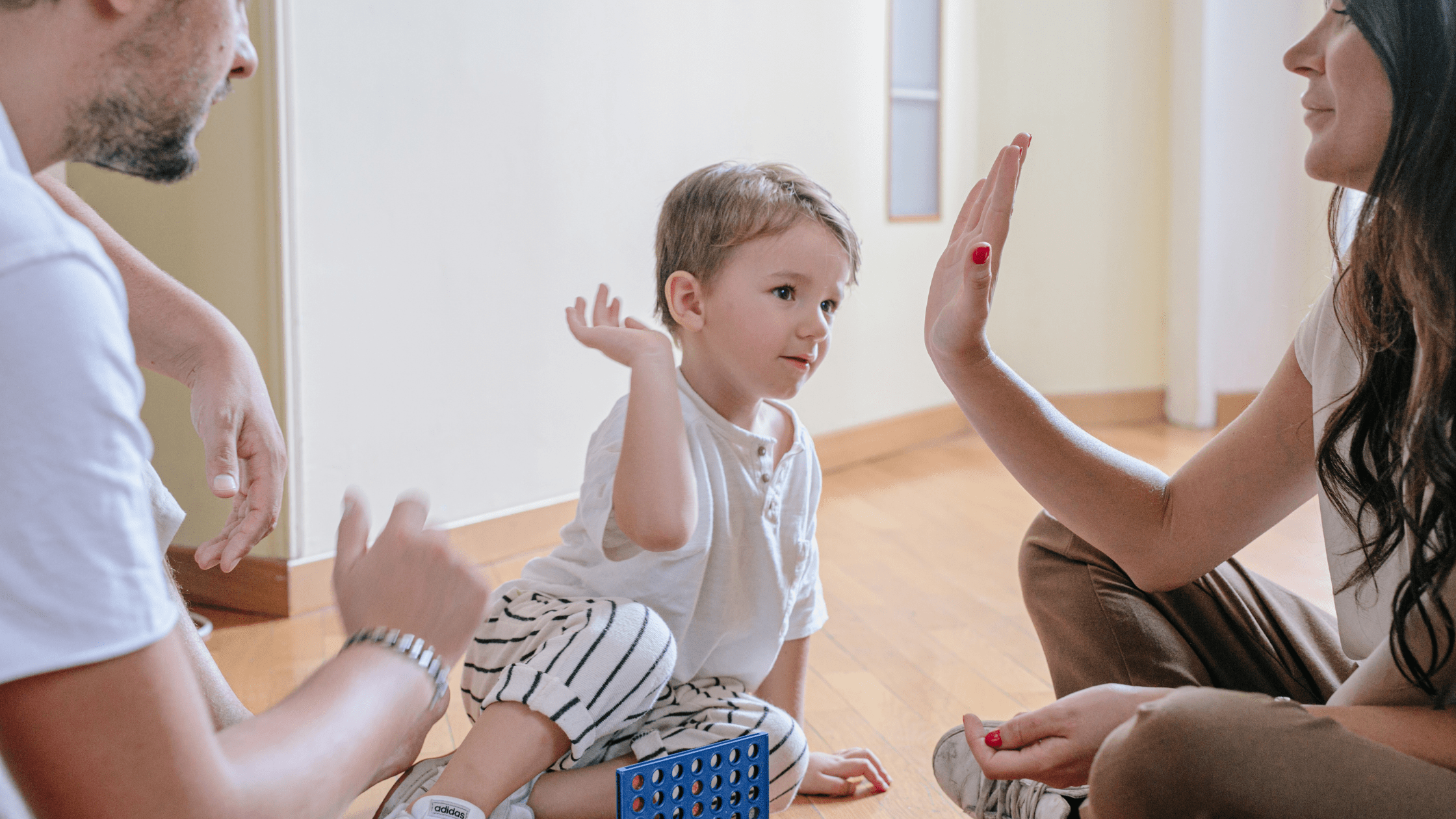 Default parent interacting with Small child sitting on the floor, giving a high-five, with another adult nearby.