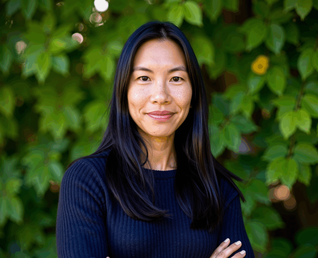 Woman with long dark hair and black sweater smiling in front of leafy green background.