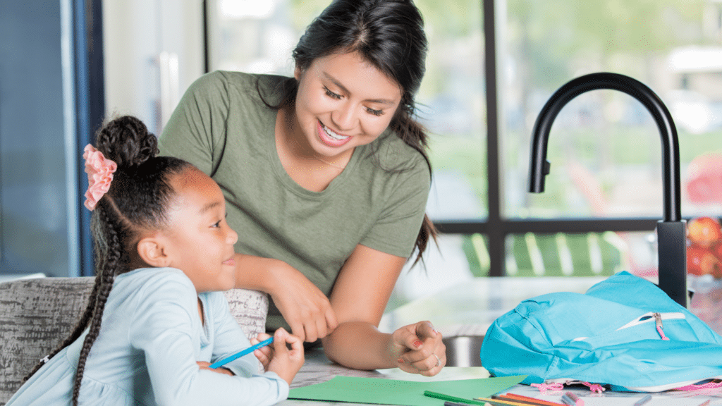 A woman smiles, helping a girl with homework at a table, surrounded by books and a blue backpack.