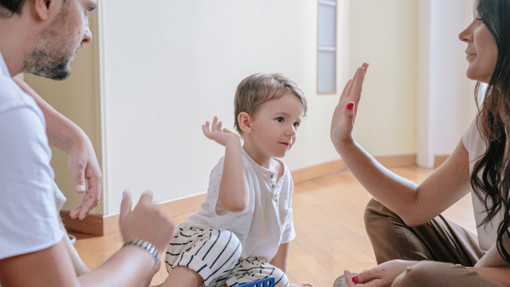 A child sitting on the floor gives a high-five to an adult, embodying fair parenting in action, while another adult watches with pride. All are dressed in casual clothes, creating a warm and nurturing atmosphere.