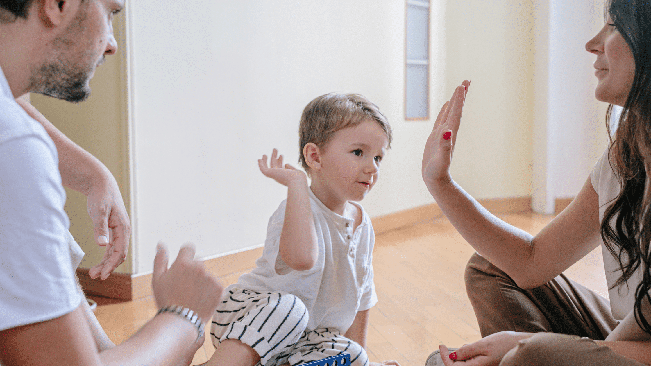 A child sitting on the floor gives a high-five to an adult, embodying fair parenting in action, while another adult watches with pride. All are dressed in casual clothes, creating a warm and nurturing atmosphere.