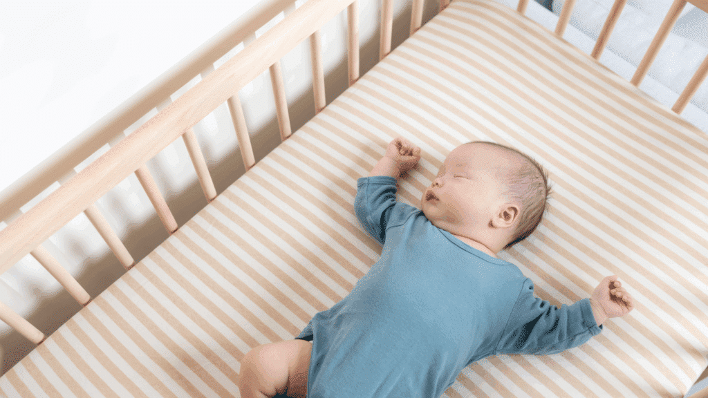 Baby in a blue onesie sleeping on a striped sheet in a wooden crib, arms stretched out.