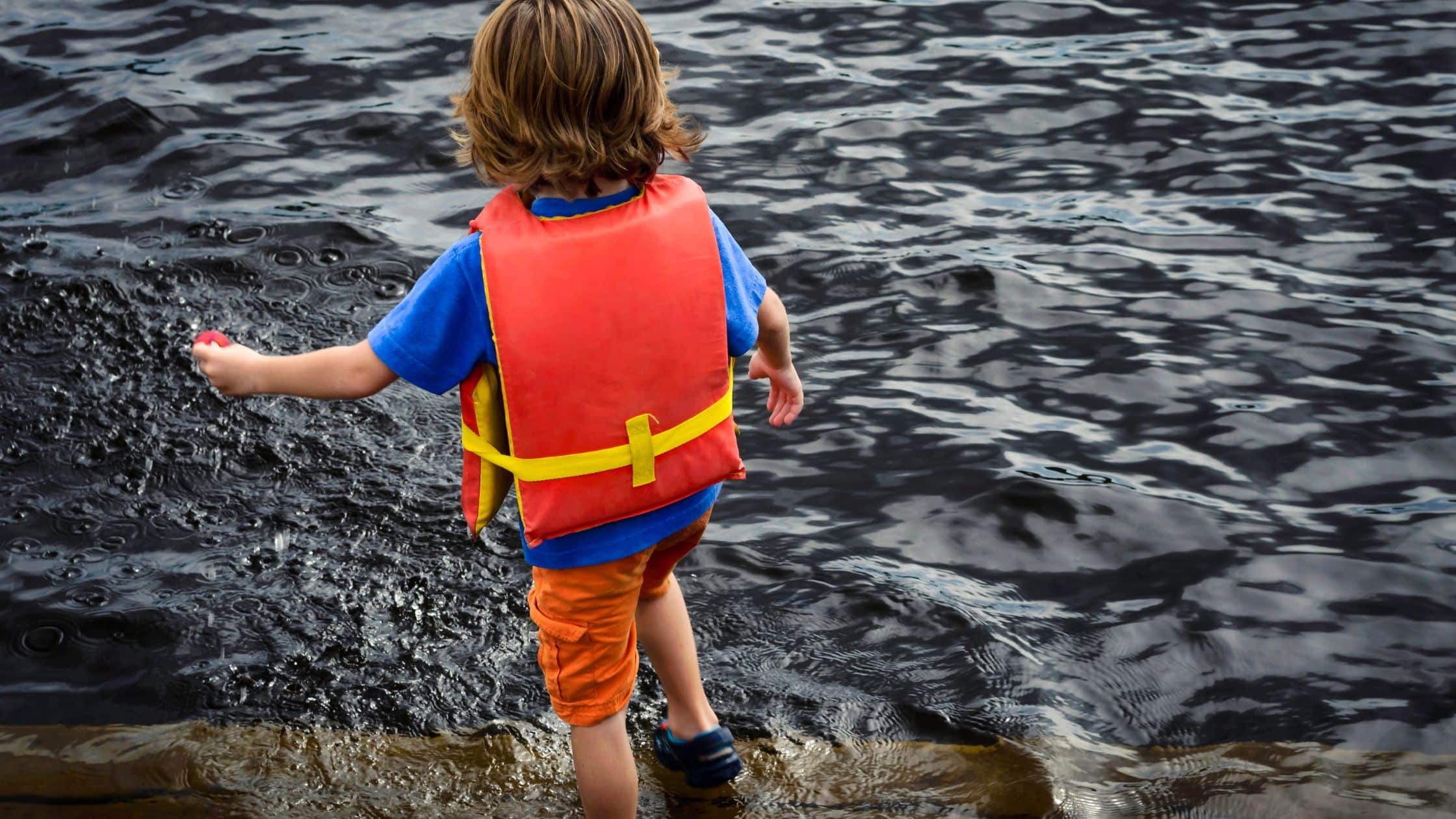 Water safety tips for kids Child wearing a red life jacket and orange shorts stands at the edge of dark, rippling water.