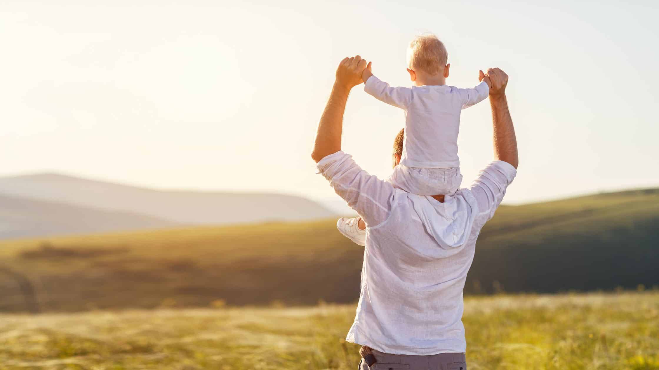 An adult carrying a child on their shoulders in a sunlit field, facing away toward hills in the distance—a classic moment that embodies the spirit of Dad Brands.