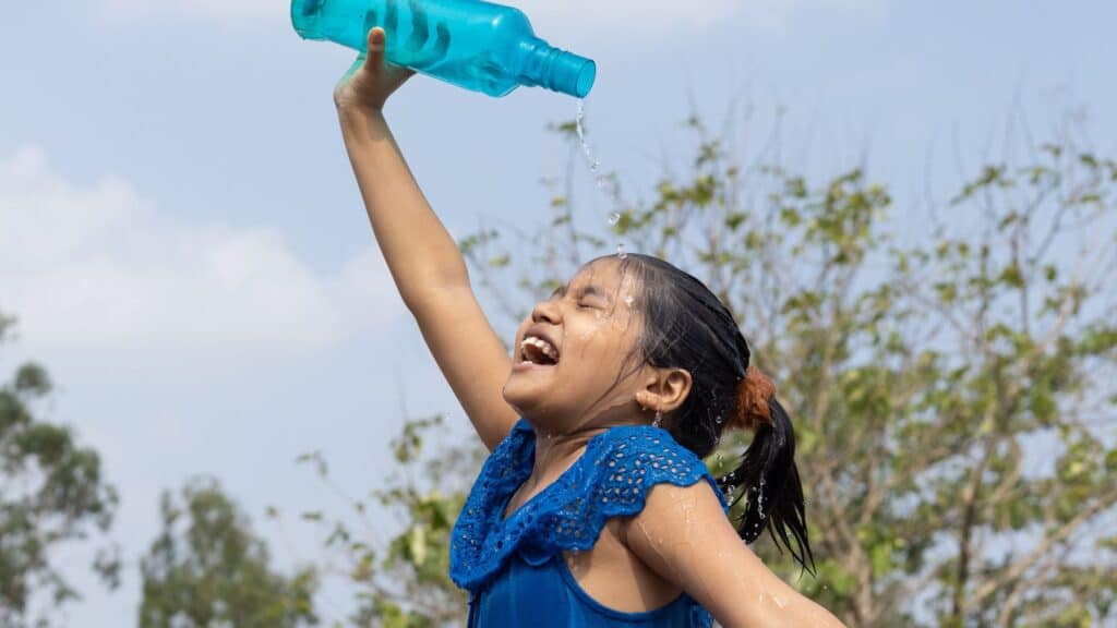 Smiling girl pours water over her head from a bottle outdoors on a sunny day, cooling off and preventing heat exhaustion.