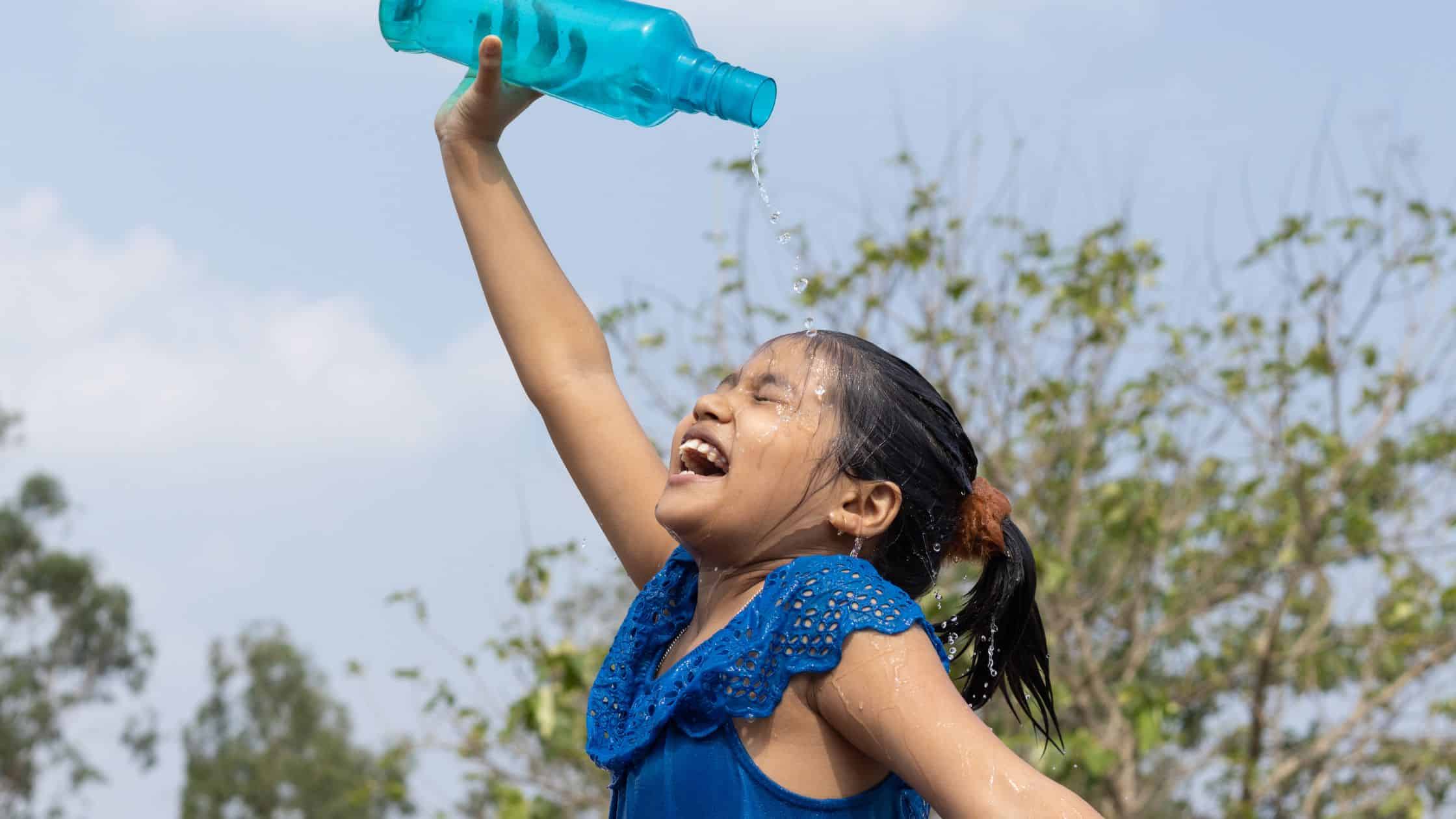 Smiling girl pours water over her head from a bottle outdoors on a sunny day, cooling off and preventing heat exhaustion.