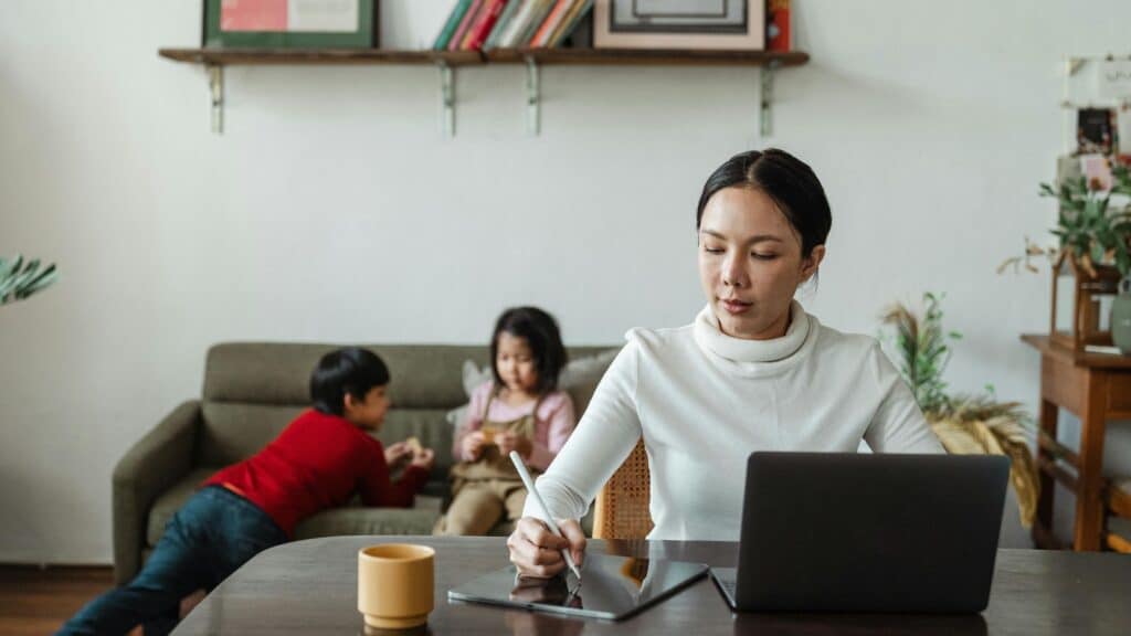 A woman manages time confetti as she works at her desk with a laptop, while two children play on a couch in the background.