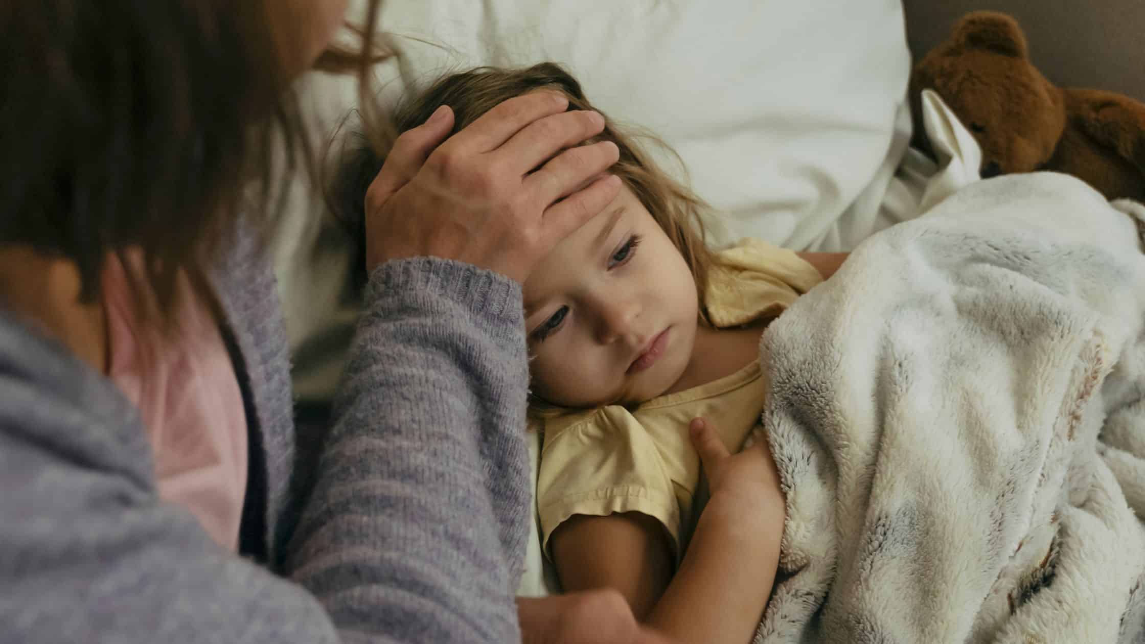 An adult feels a sick child's forehead as she lies in bed under a blanket, looking unwell, reflecting those moments that often lead to seeking pediatrician advice for common pediatric questions.