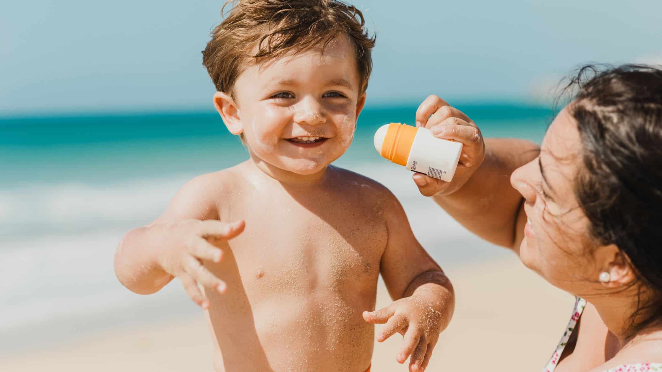 Smiling toddler on the beach as an adult applies sunscreen to his face.