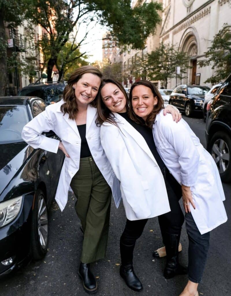 Three smiling women in white lab coats pose together on a city street lined with cars and trees.