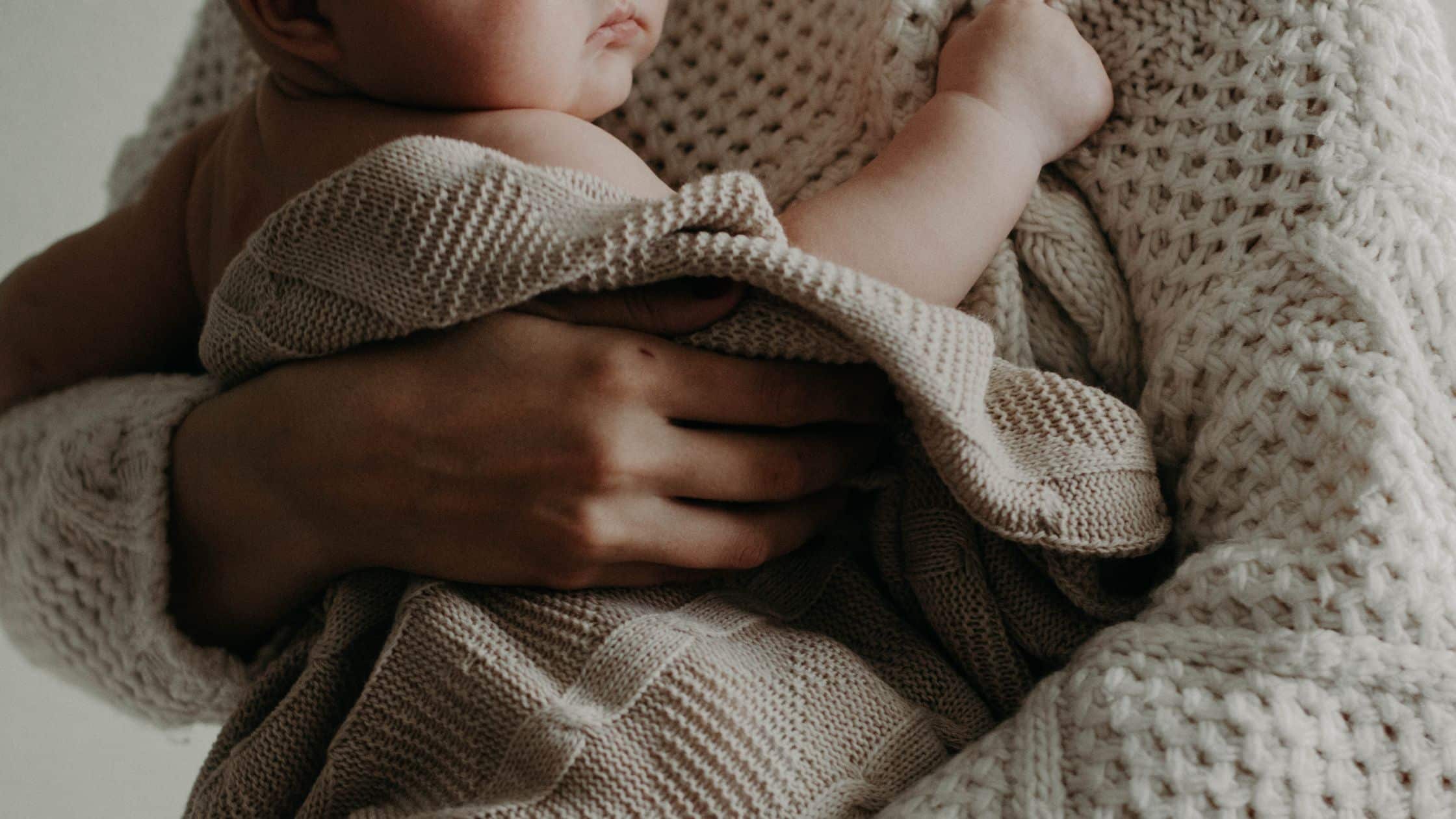 A baby wrapped in a knitted blanket is held closely by an adult wearing a chunky knit sweater.