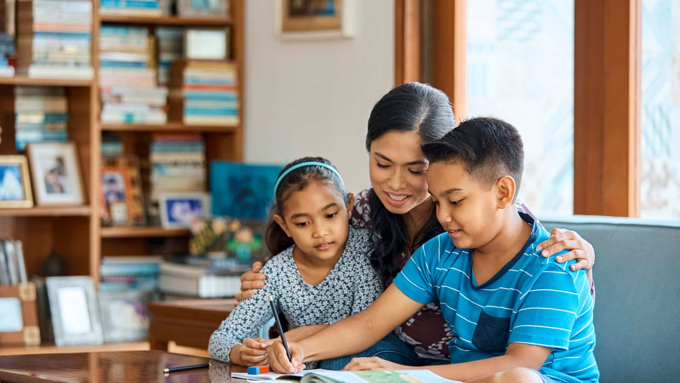 A woman helps two children with homework at a table in a cozy, book-filled living room—perfect inspiration for transitioning your nanny to a family assistant.