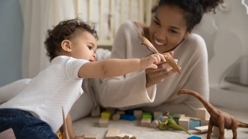 Mother and young child playing with toy dinosaurs and wooden blocks on the floor together.
