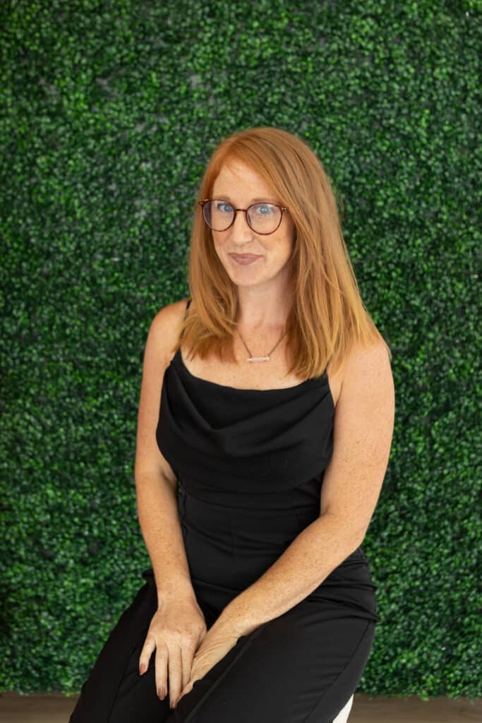 Woman with red hair and glasses sits in front of a green leafy wall, wearing a black sleeveless outfit.
